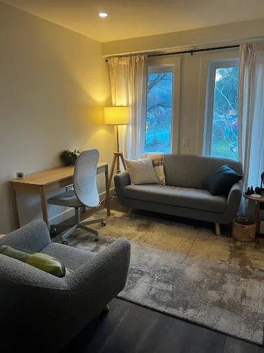 Living room with a gray sofa, a gray armchair, a wooden desk with a white chair, a floor lamp, and large windows with white curtains, during evening.