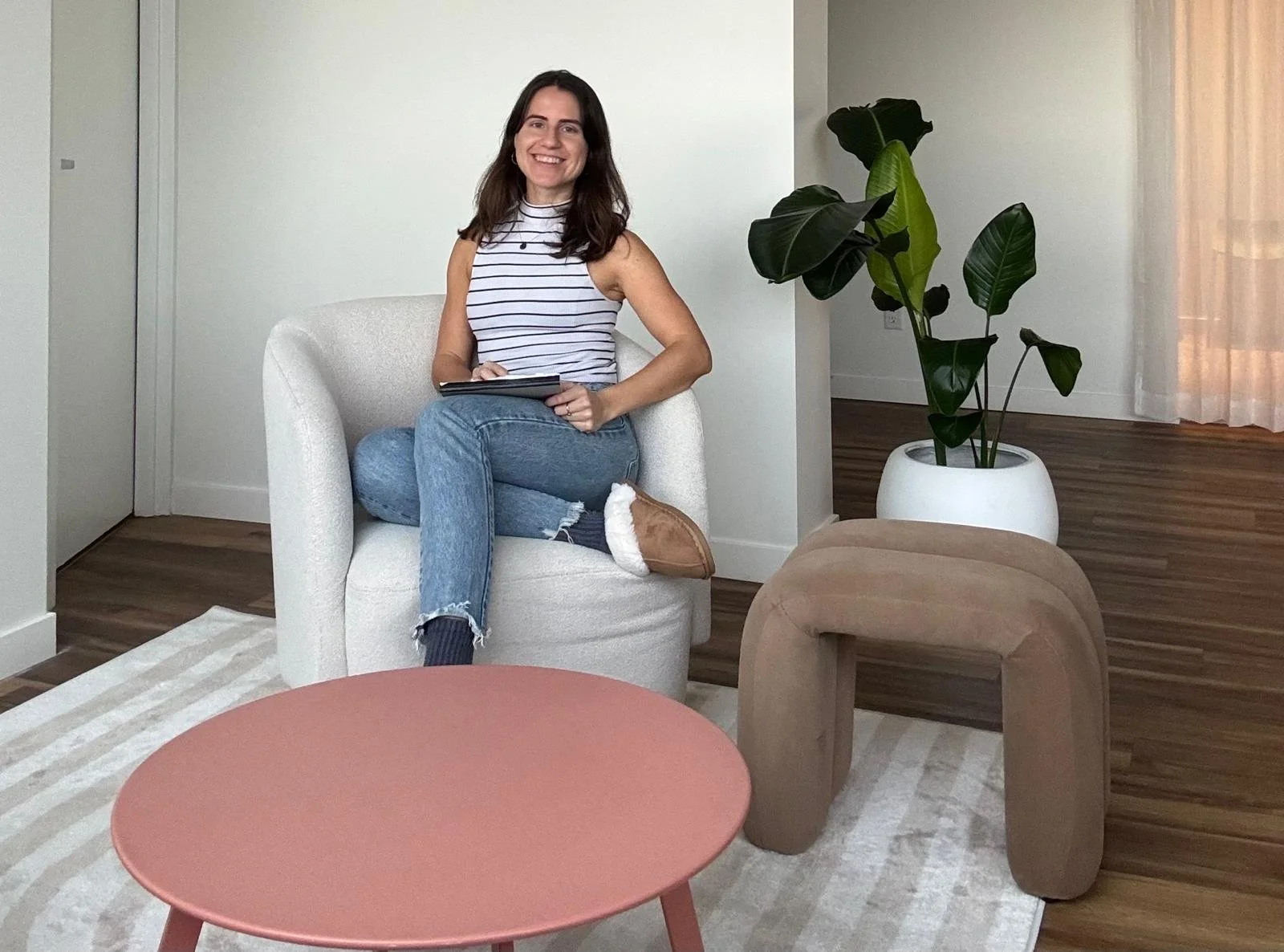 A woman with dark hair, wearing a striped sleeveless top and jeans, sitting in a cozy, modern living room chair, smiling, with a notebook in her lap. The living room features a patterned rug, a round pink coffee table, a large potted plant, and sheer curtains.
