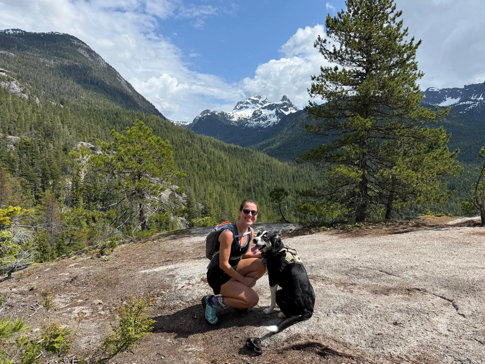 A woman smiling and squatting next to a dog on a mountain trail with a forest and snow-capped peaks in the background on a partly cloudy day.