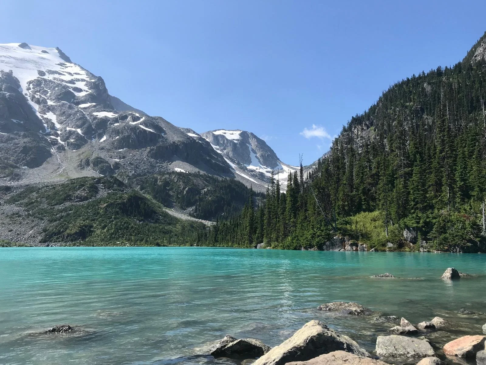 Mountain lake with turquoise water, surrounded by evergreen trees and snow-capped peaks under a clear blue sky.