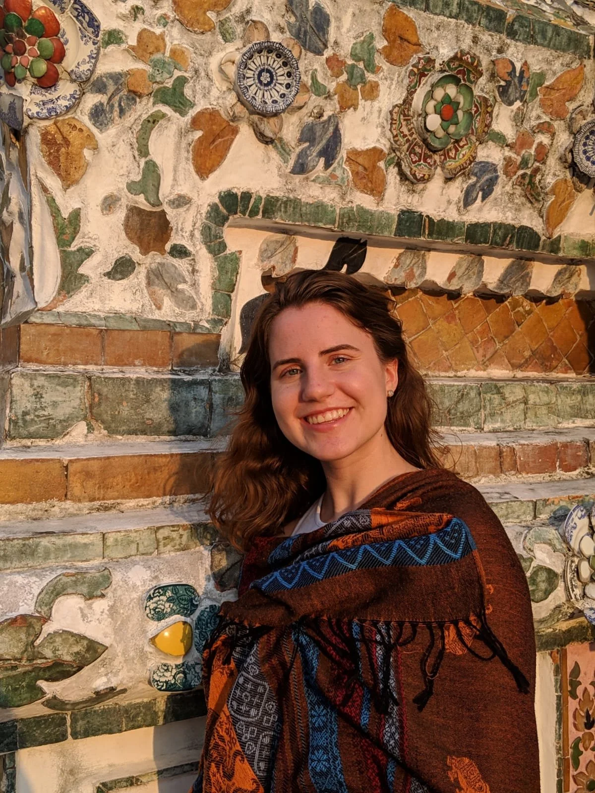 A smiling woman with wavy brown hair and a colorful blanket over her shoulders, standing in front of an intricate stone wall decorated with ceramic tiles and mosaics.