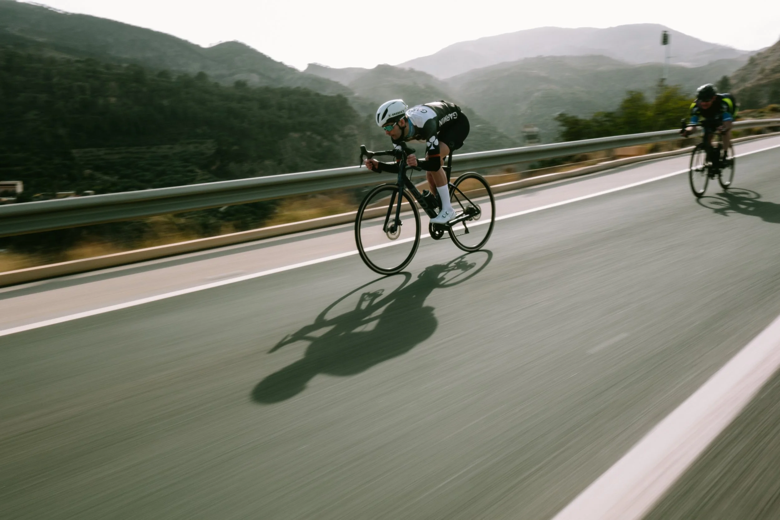 Two cyclists riding on a highway with mountains in the background, wearing helmets and sports gear.