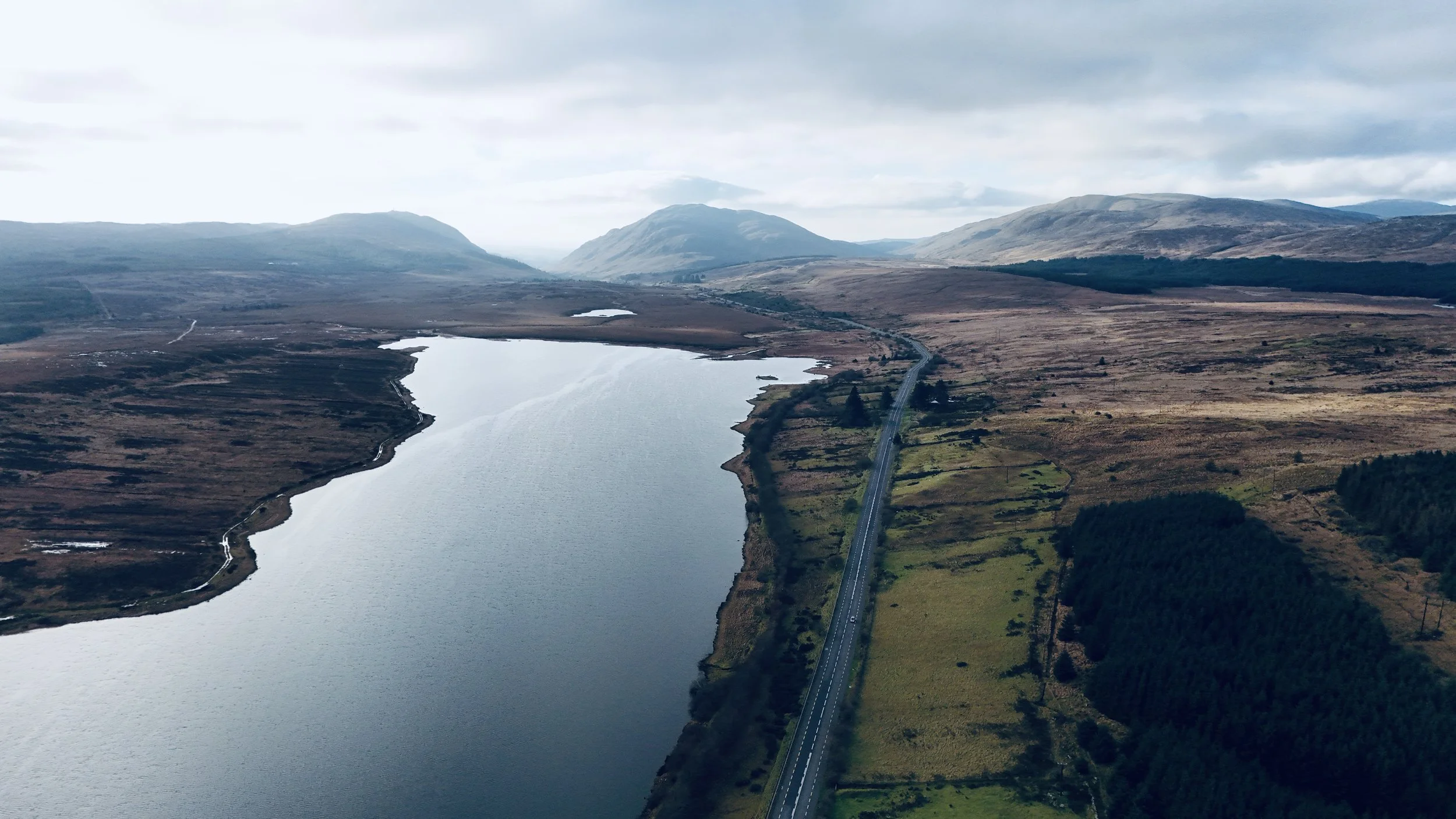 Aerial view of a narrow road running alongside a large lake, with expansive plains and hills in the background under cloudy sky.