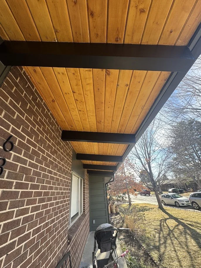 View of a house porch with a brick wall, window, and wooden ceiling with black support beams, sunny day, parked cars, trees, and grass in the background.