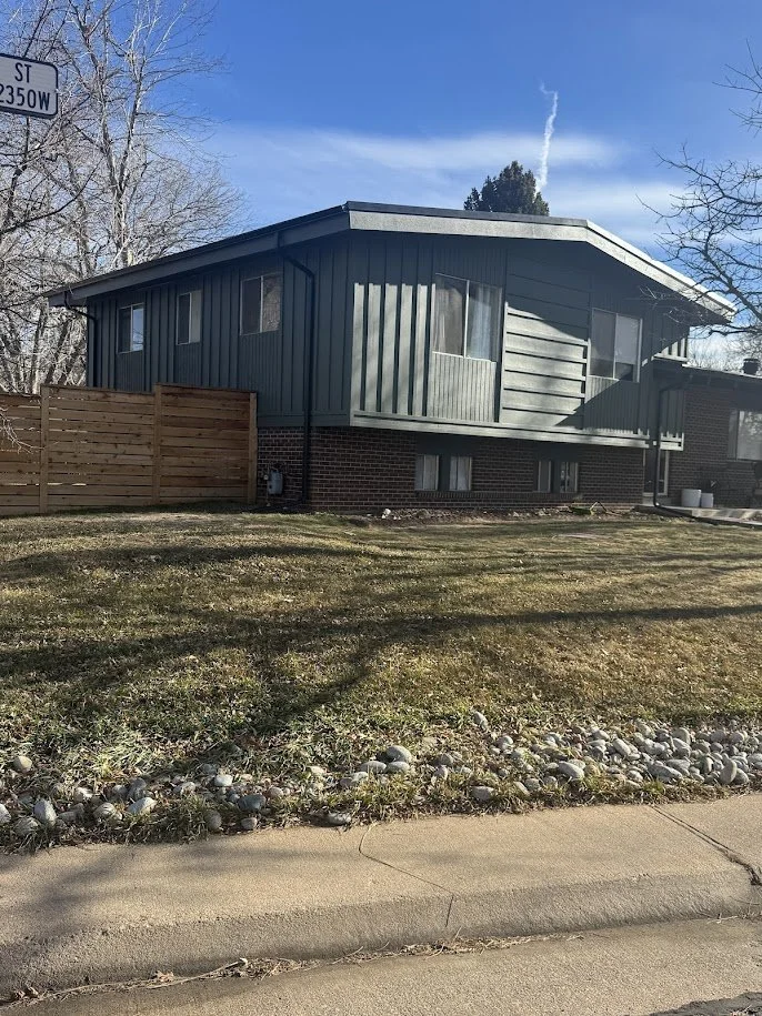 A two-story house with blue siding and a brick foundation, surrounded by a wooden fence on one side and a small front yard with patches of grass and rocks, under a clear blue sky.