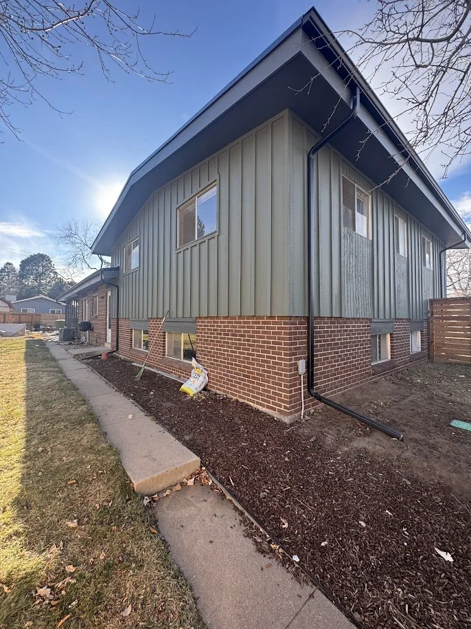 Side view of a two-story house with brick foundation and green siding, with a sloped roof, black gutter, and exposed garden bed along the walkway.