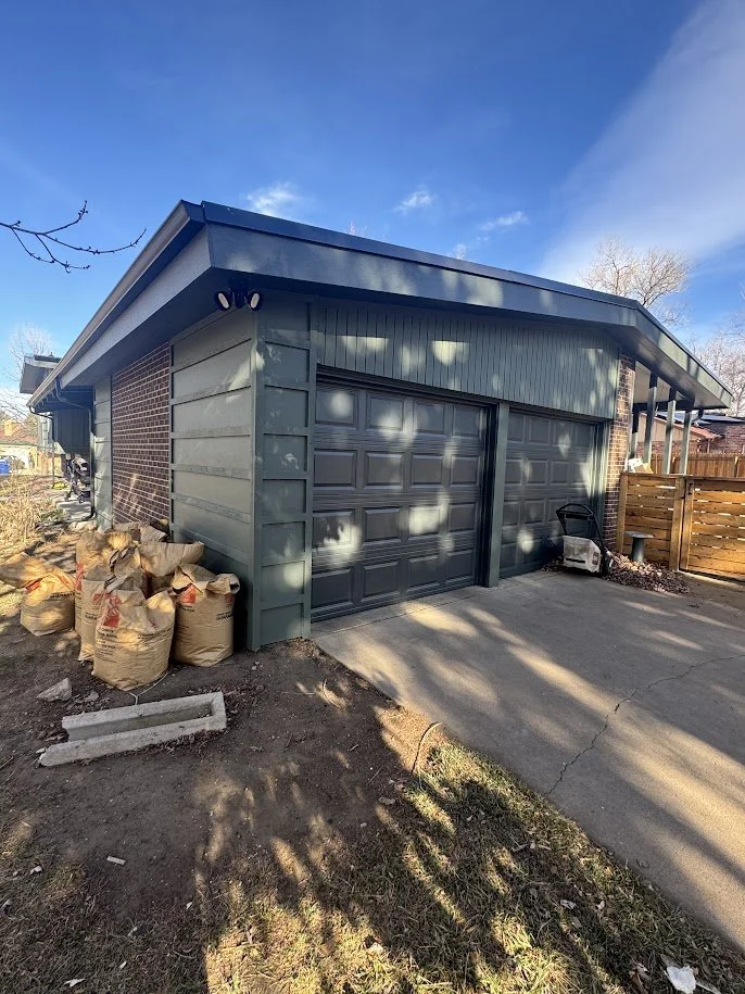 Newly constructed garage with a gray roll-up door, brick and siding exterior, and construction materials on the side, under a blue sky.