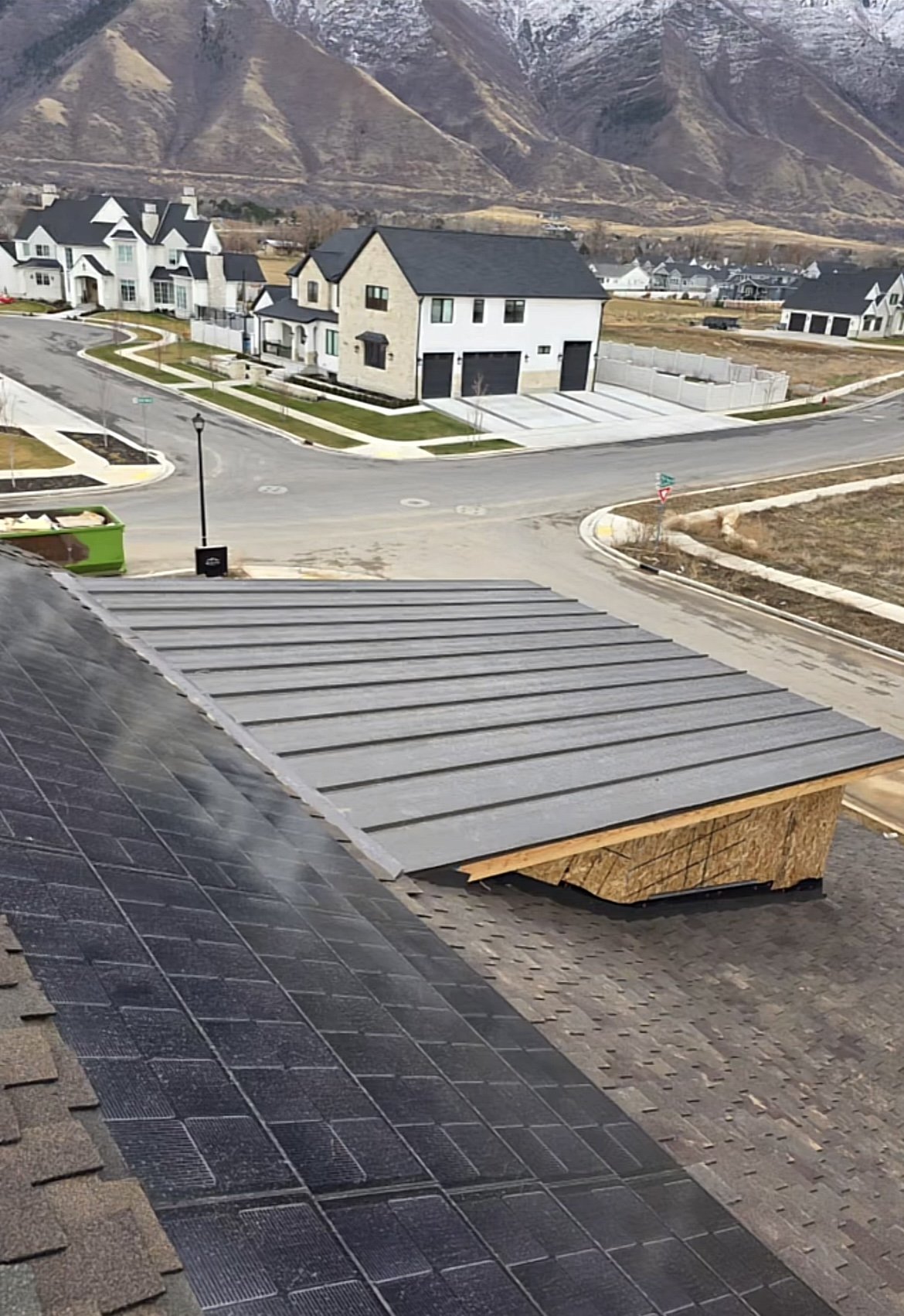 A view from a rooftop showing solar panels and part of a roof under construction in a suburban neighborhood. In the background, there are finished houses, streets, and mountains.
