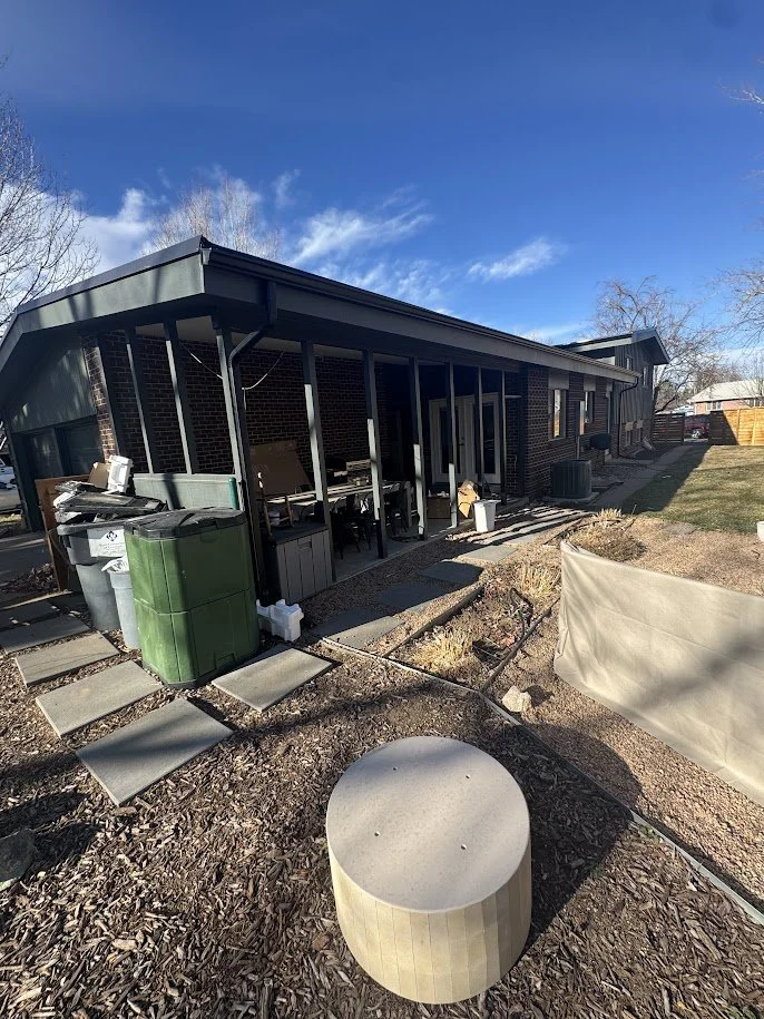 Backyard of a brick house with a covered patio, outdoor furniture, and trash bins, with a wooden fence and a fence with a privacy screen in the distance.