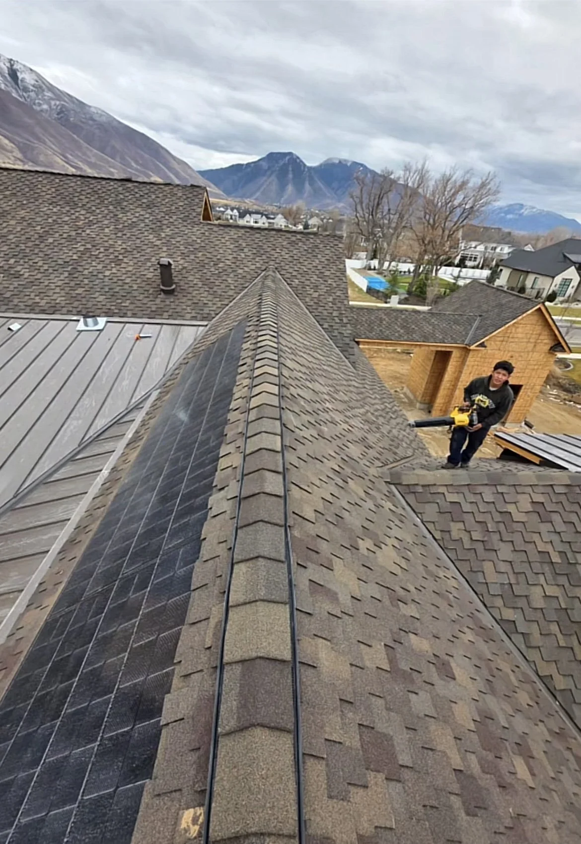 A person working on a sloped brick roof with solar panels, holding a leaf blower, with mountains and residential houses in the background.