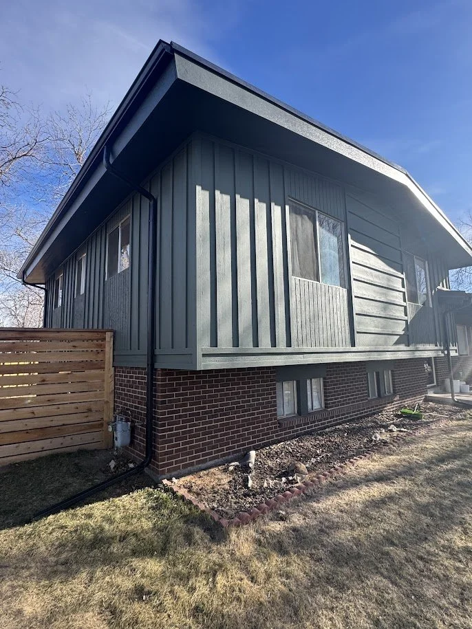 Backside of a house with dark green vertical siding, brick foundation, and small basement windows. There is a wooden privacy fence and a grassy yard.