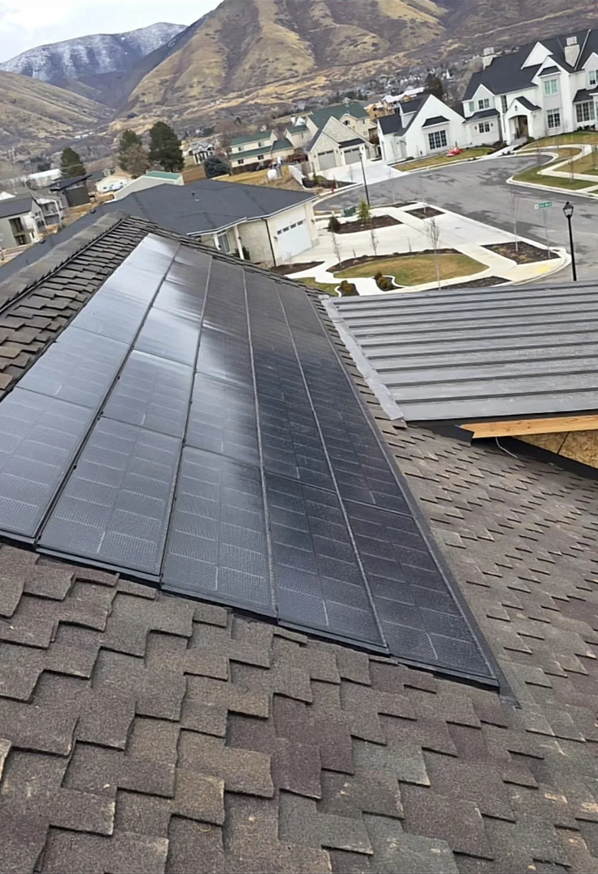 Residential rooftop with solar panels overlooking a suburban neighborhood with houses, street, sidewalks, lamp posts, landscaped yards, and mountains in the background.