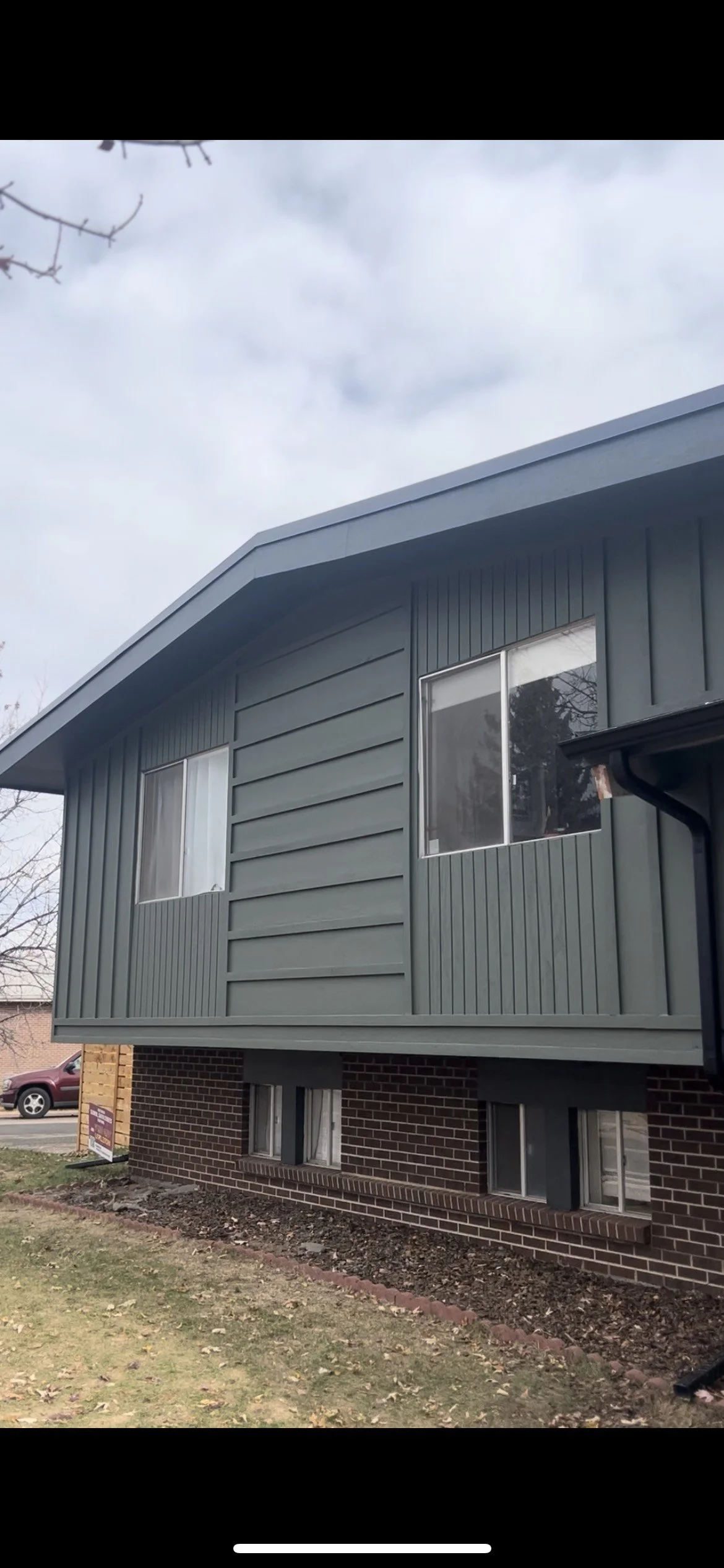 View of a two-story house with a brick lower level and a green vinyl siding upper level, featuring two windows on each level, a sloped roof, and a small front yard.