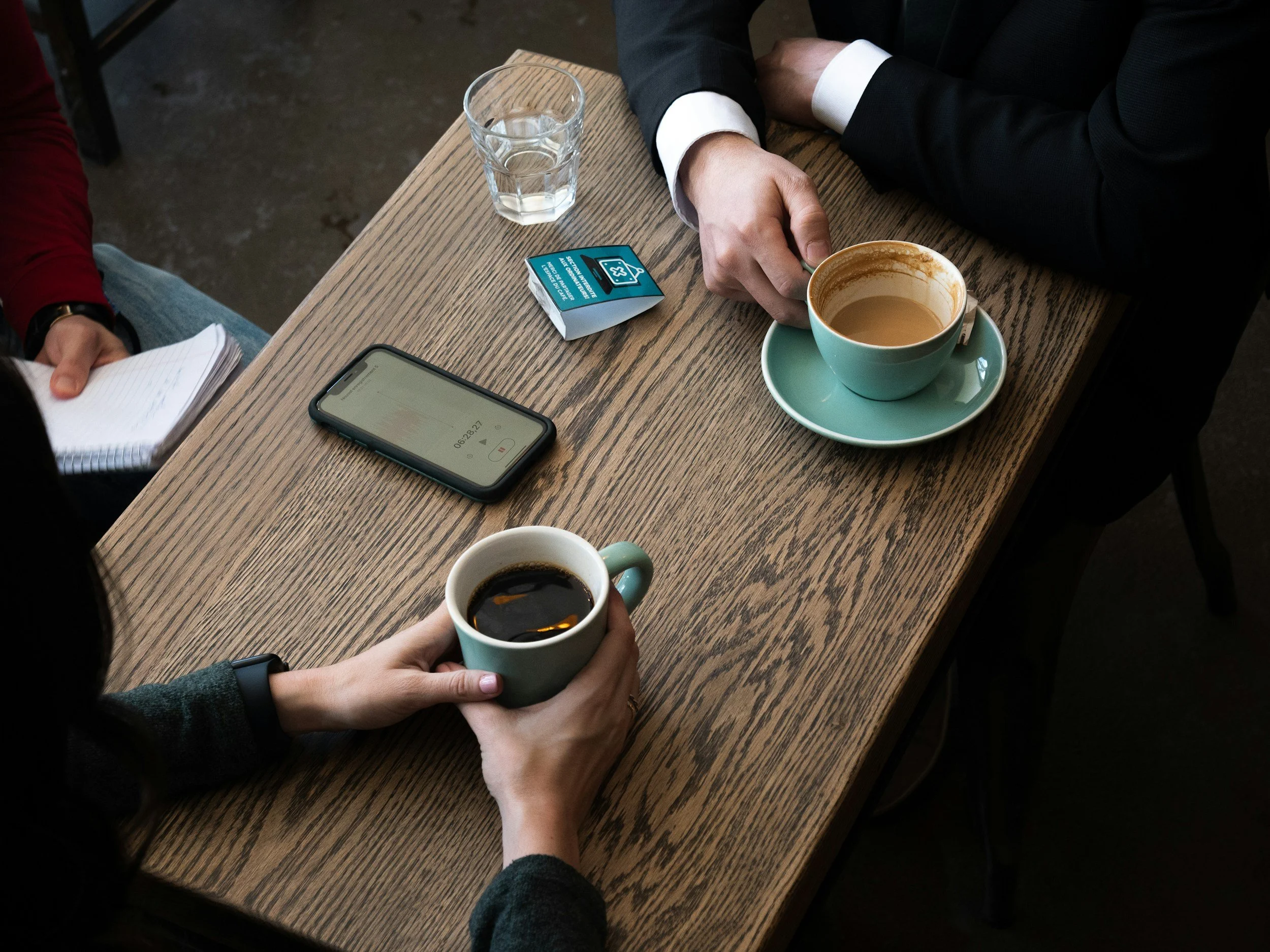 Two people are sitting at a wooden table with coffee cups, a glass of water, a smartphone, a small pack of sugar, and a notepad. One person is holding a dark coffee mug, and the other is reaching for a light green coffee mug with their hand.