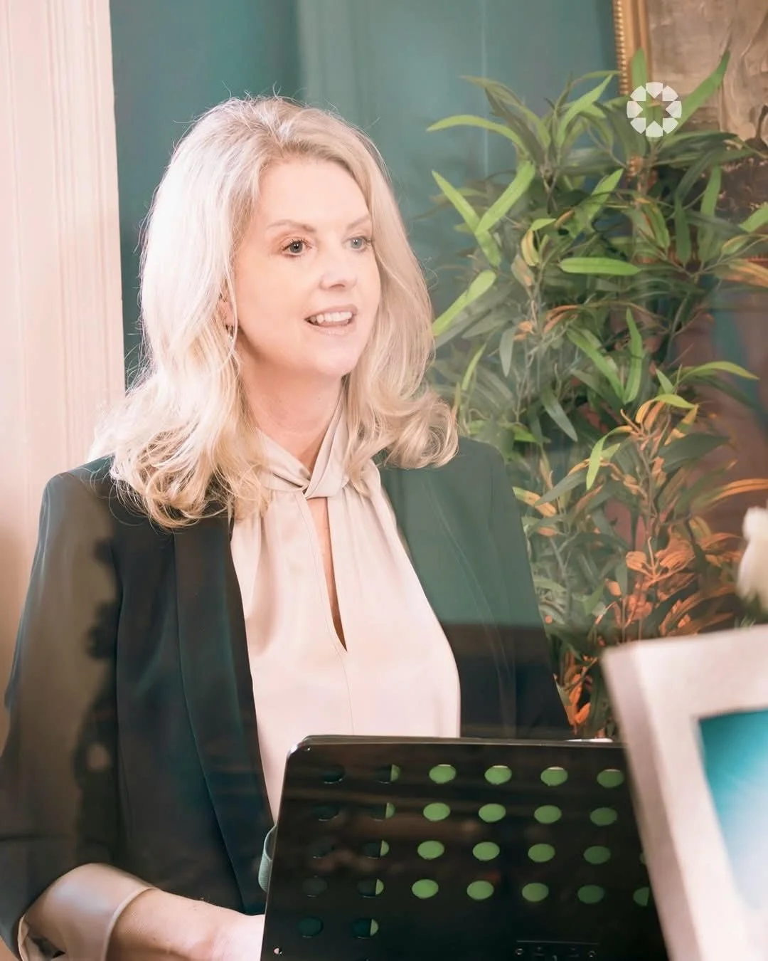A woman with blonde hair wearing a beige blouse and a dark blazer, sitting at a desk with a black music stand in front of her. There are green plants in the background.