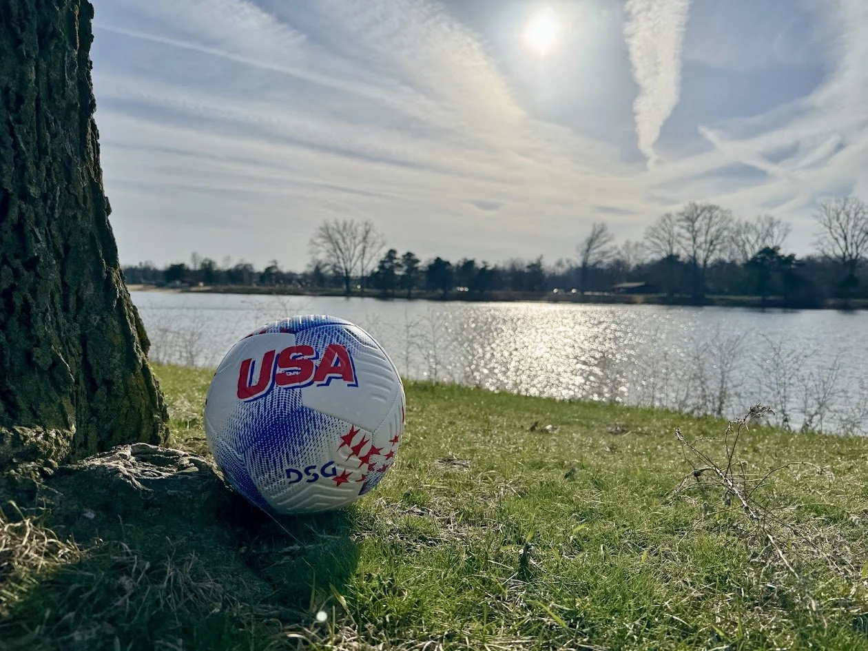 A soccer ball with USA markings resting on the grass near a tree by a lake during daytime with a partly cloudy sky.