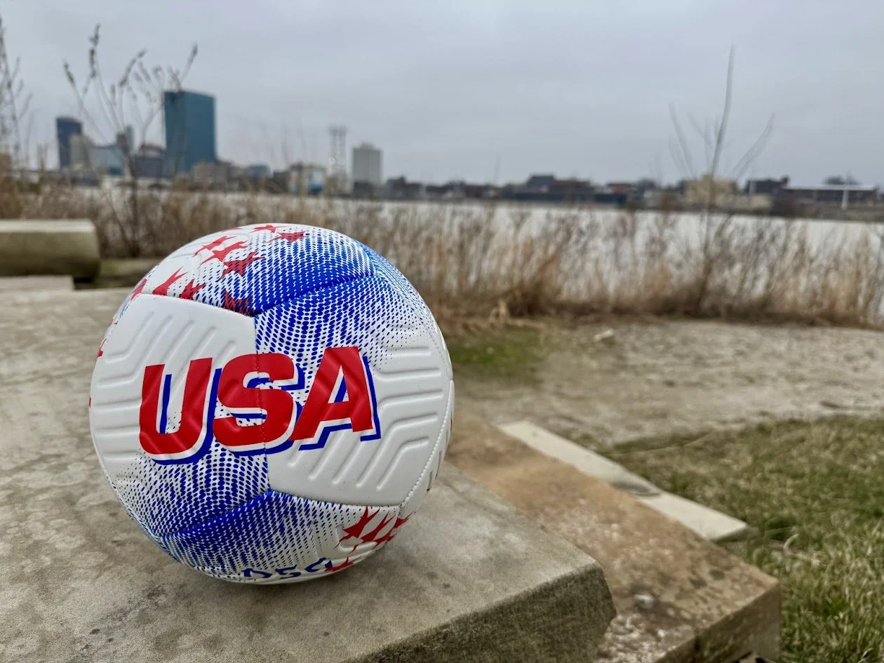 A white soccer ball with red, white, and blue design and the text "USA" in red letters, resting on a stone surface outdoors with a city skyline and overcast sky in the background.