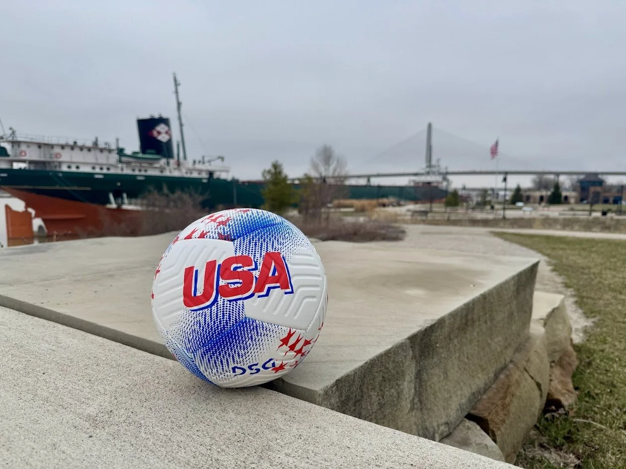 A soccer ball with USA colors and text sitting on a stone ledge near a harbor with ships and a bridge in the background.