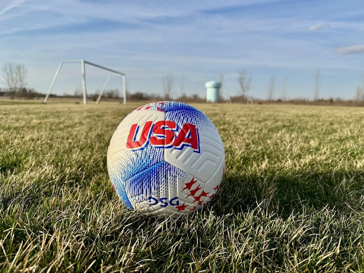 Soccer ball with USA and PSC logos on grass field with goalposts and a water tower in the background under blue sky.