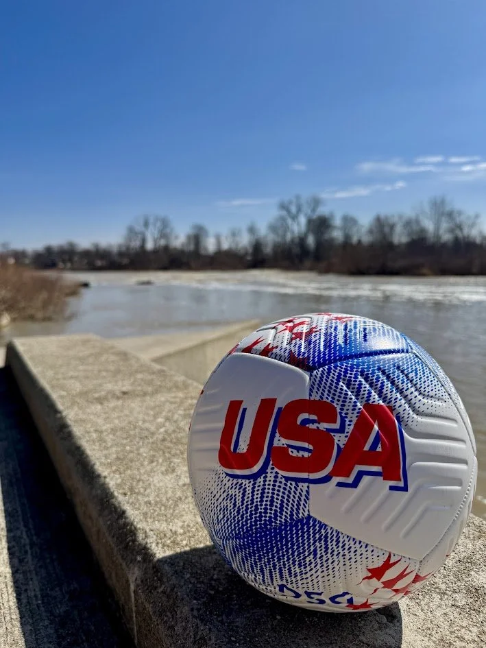 A soccer ball with USA branding resting on a concrete ledge by a river on a sunny day with a clear blue sky.