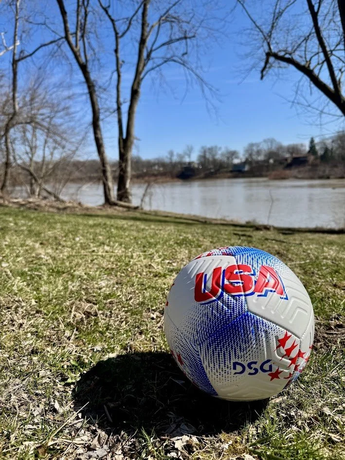 A soccer ball with 'USA' and 'DSG' printed on it, sitting on grass near a river, with leafless trees and a clear blue sky in the background.