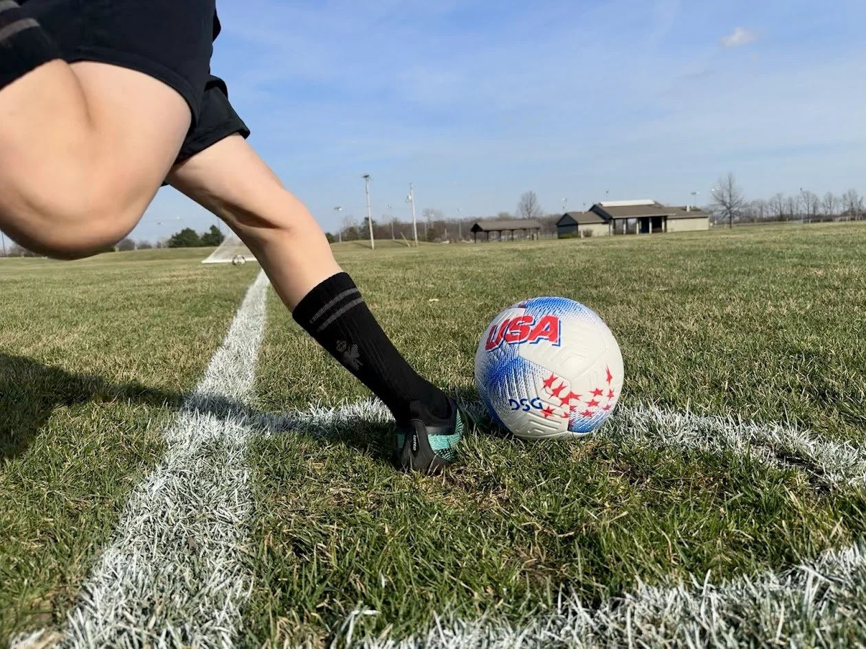 A soccer player wearing black shorts, black socks, and cleats is preparing to kick a labeled USA soccer ball on a grassy field with white boundary lines, with buildings and trees in the background.
