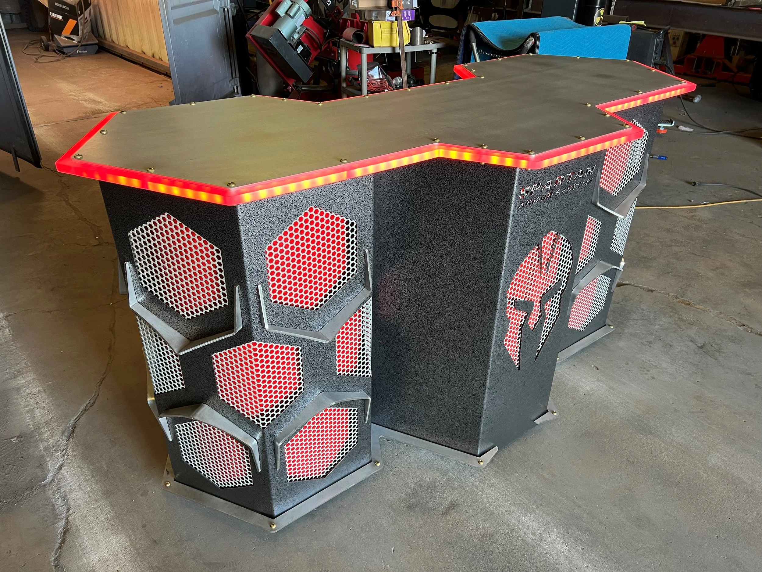 A custom gaming desk with a hexagonal pattern on its sides, black textured finish, and red LED lighting around the edge, situated on a concrete floor in a workshop or garage setting.