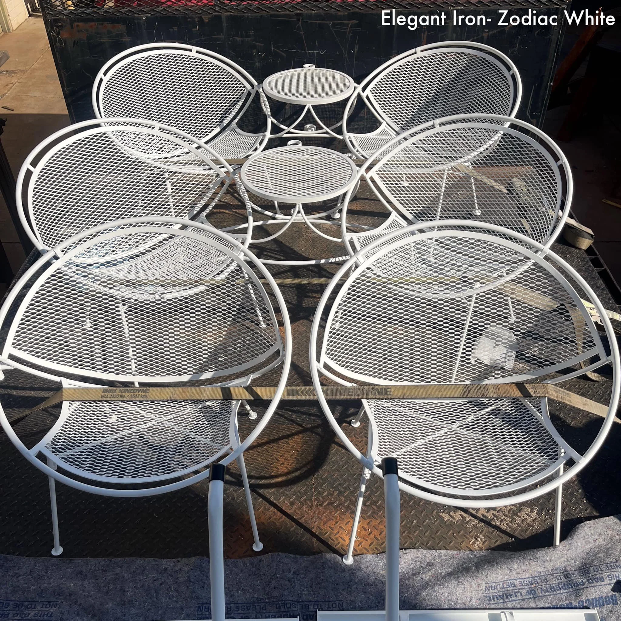 A white metal outdoor table and six matching chairs with mesh seats and backrests, arranged around the table on a workshop or storage area floor.