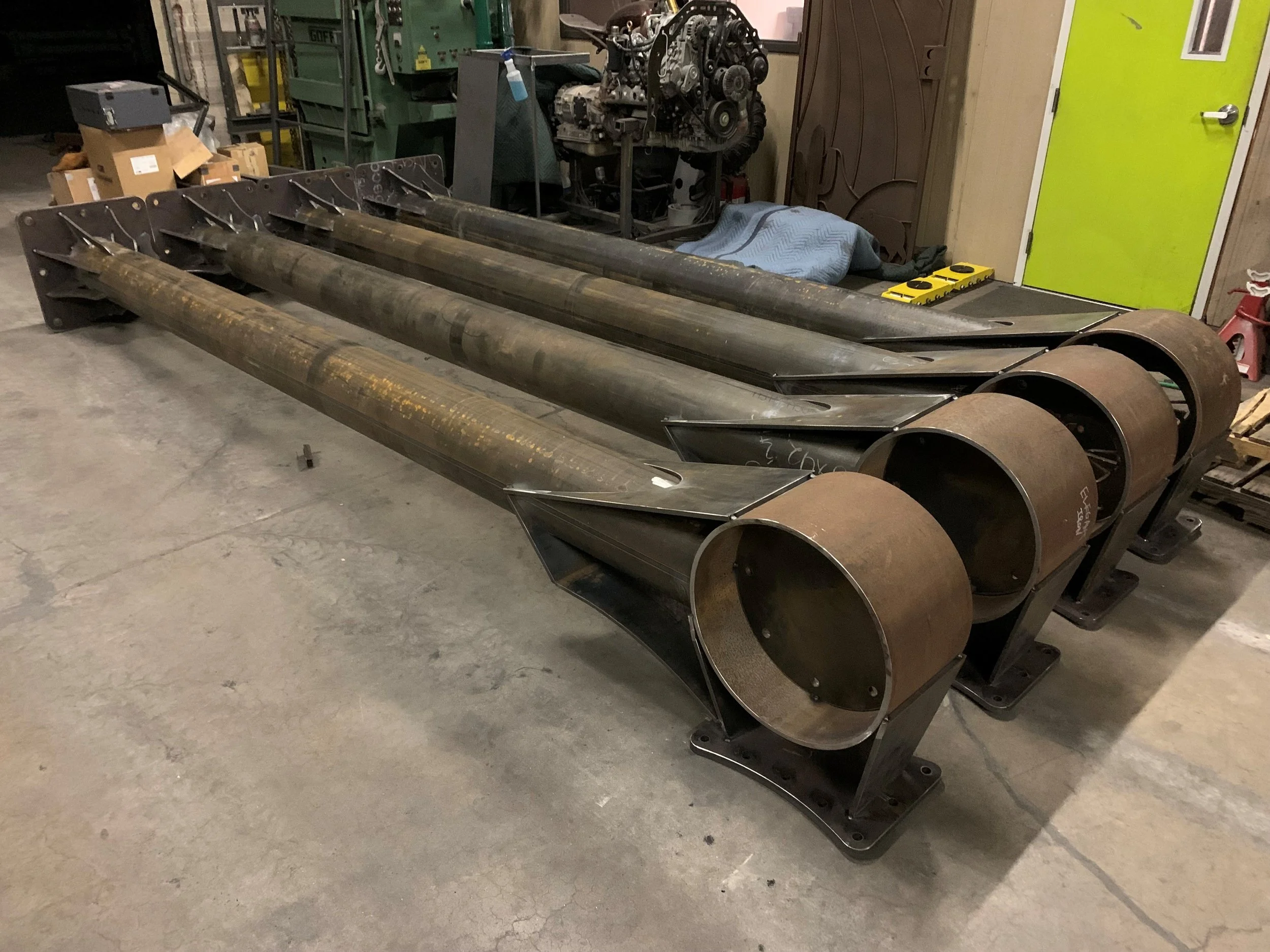 Three large steel pipes with flanges on one end lying on a workshop floor, with various tools and machinery in the background.