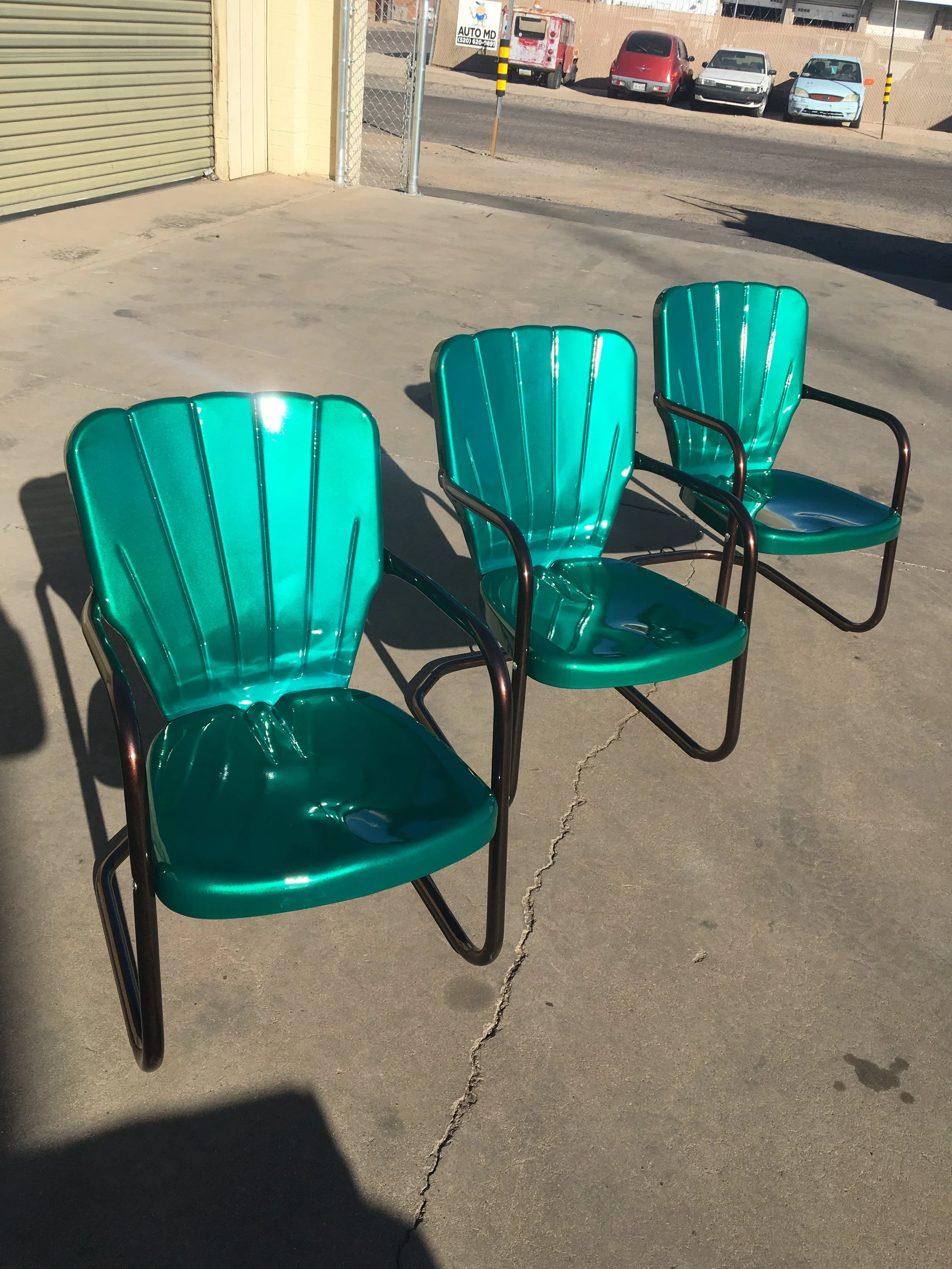 Three green metal chairs with black arms placed on a concrete sidewalk outdoors, casting shadows in sunlight.