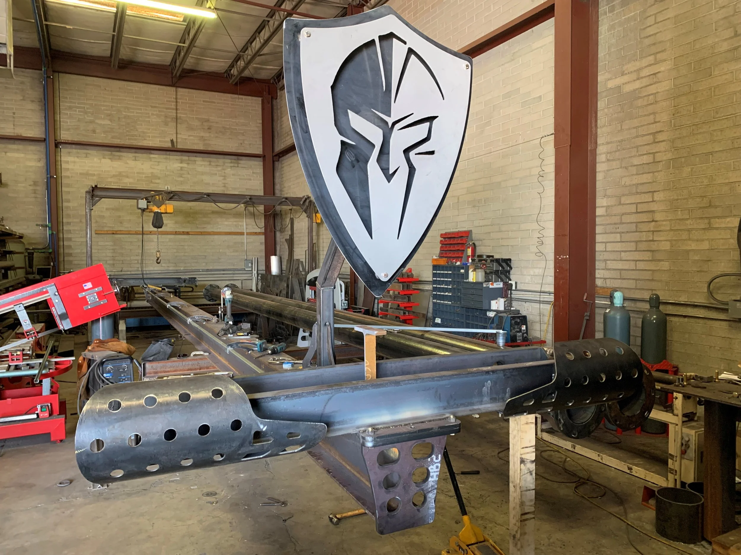 Metallic frame of a custom-made motorcycle with a shield featuring a stylized eagle emblem on the front, in a workshop with tools and equipment.