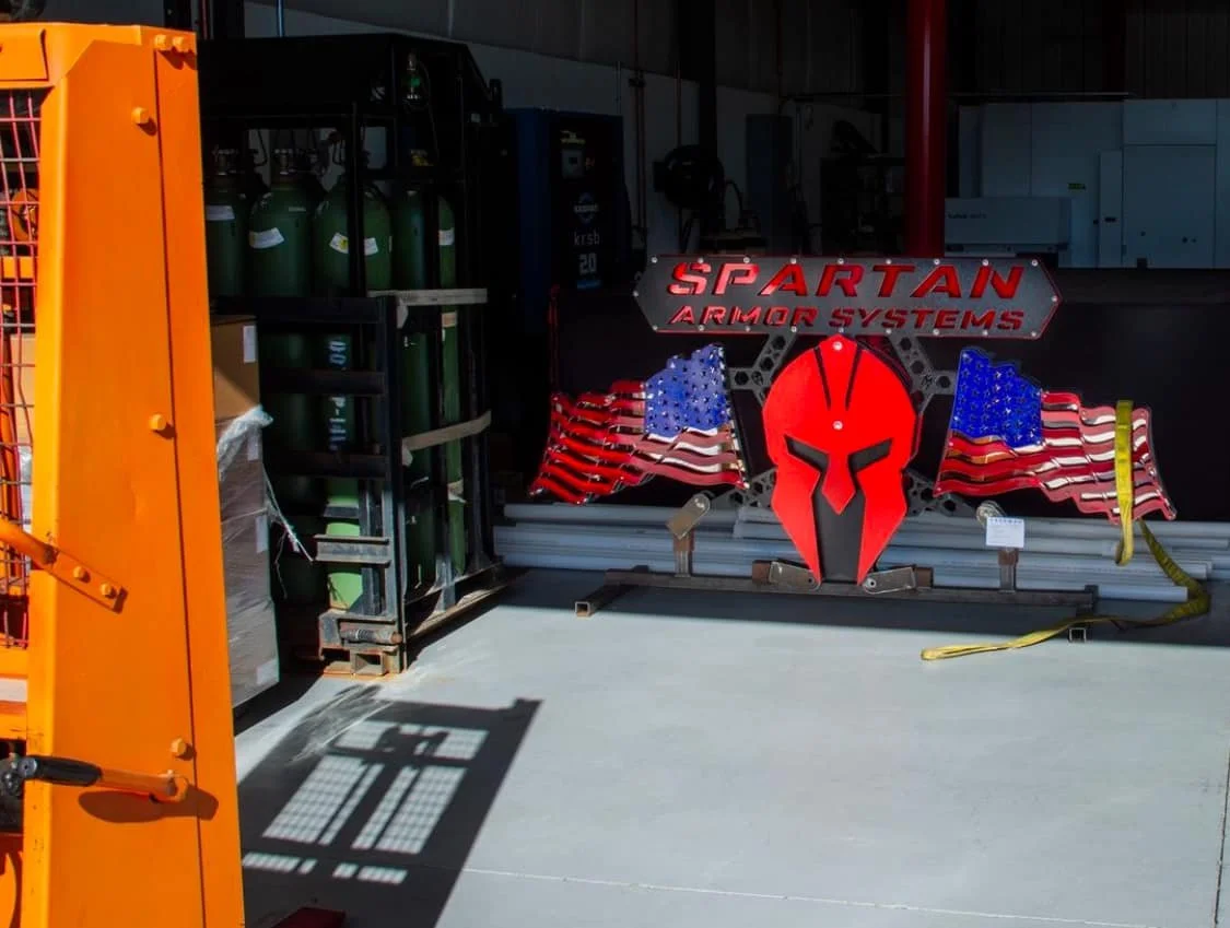 Display of Spartan Armor Systems logo with American flags and a red Spartan helmet in an industrial setting, with gas cylinders and equipment in the background.