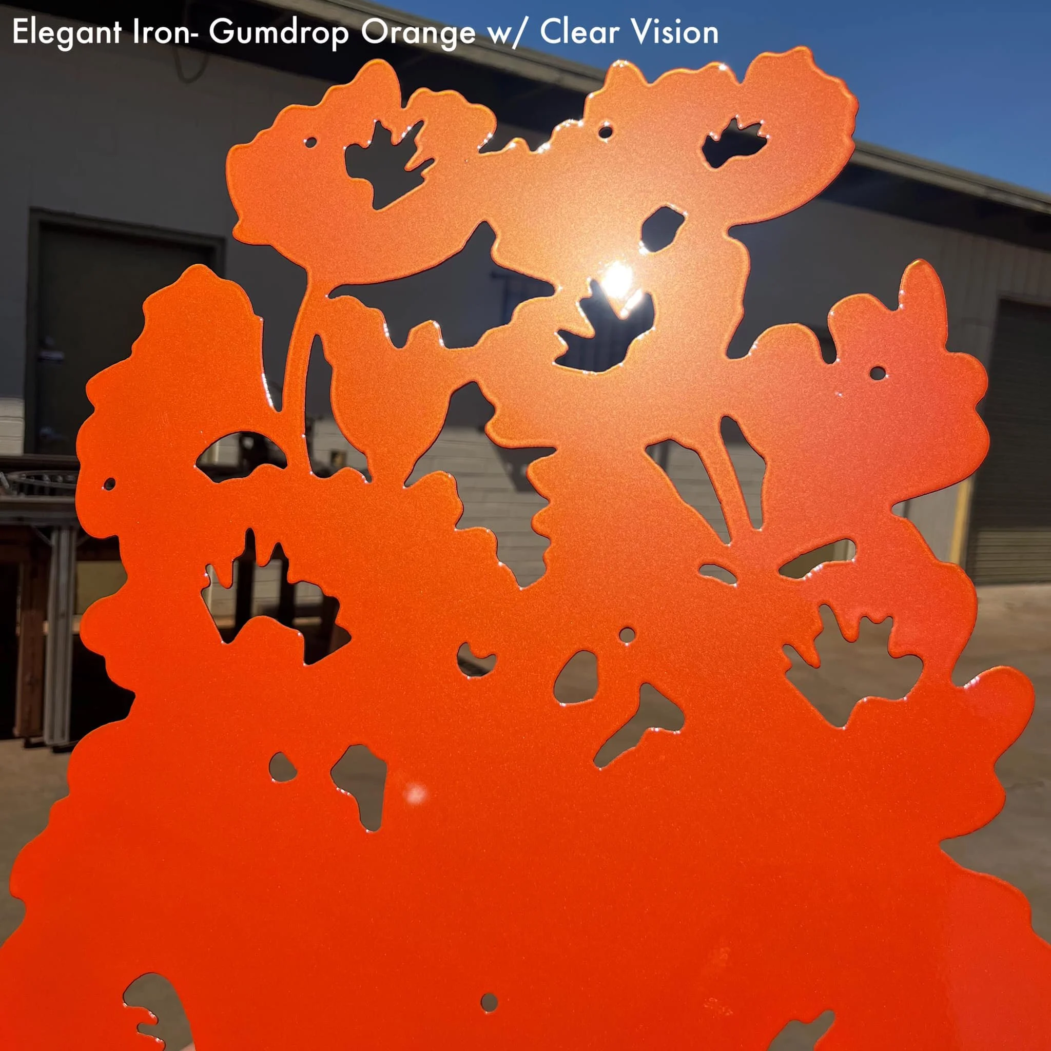Close-up of an orange metal garden decoration with butterfly-shaped cutouts, outdoors with a building and blue sky in the background.