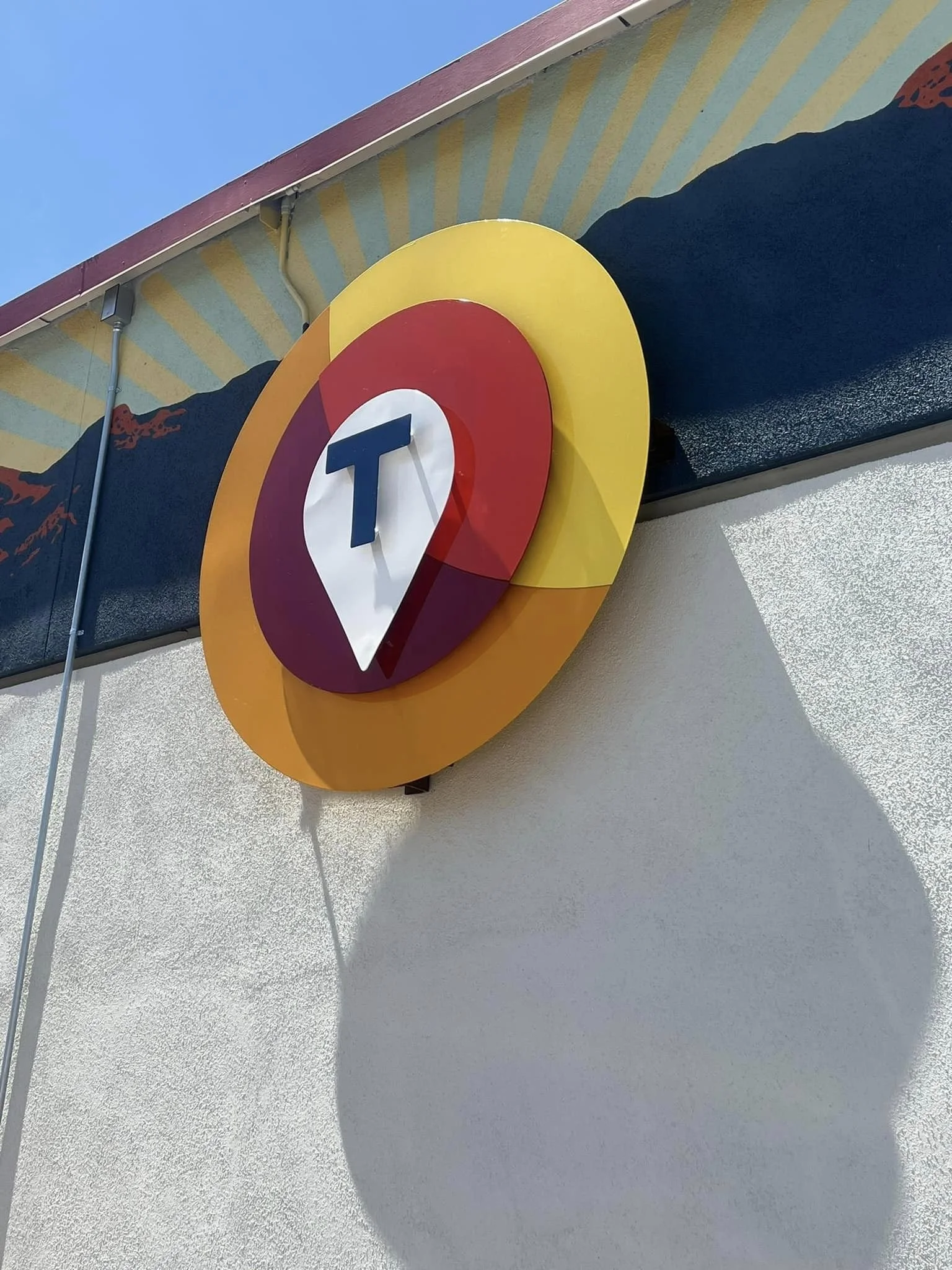 Colorful Circle Sign with a white map pin icon and a dark blue letter 'T' in the center, mounted on a white wall outside a building.