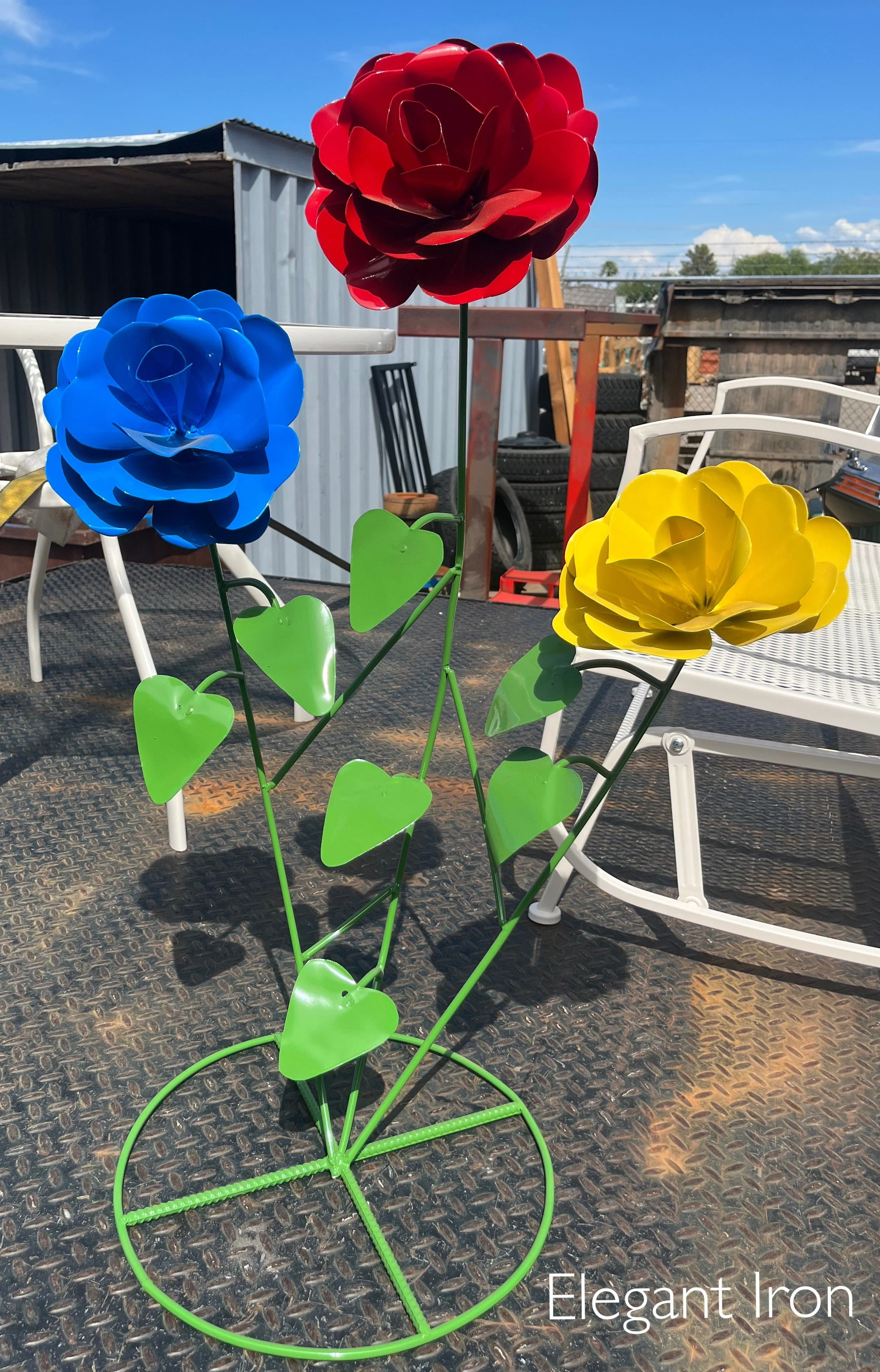 Colorful metal flower sculpture with large red, blue, and yellow flowers and green leaves, outdoors on a metal surface with chairs and a shed in the background.