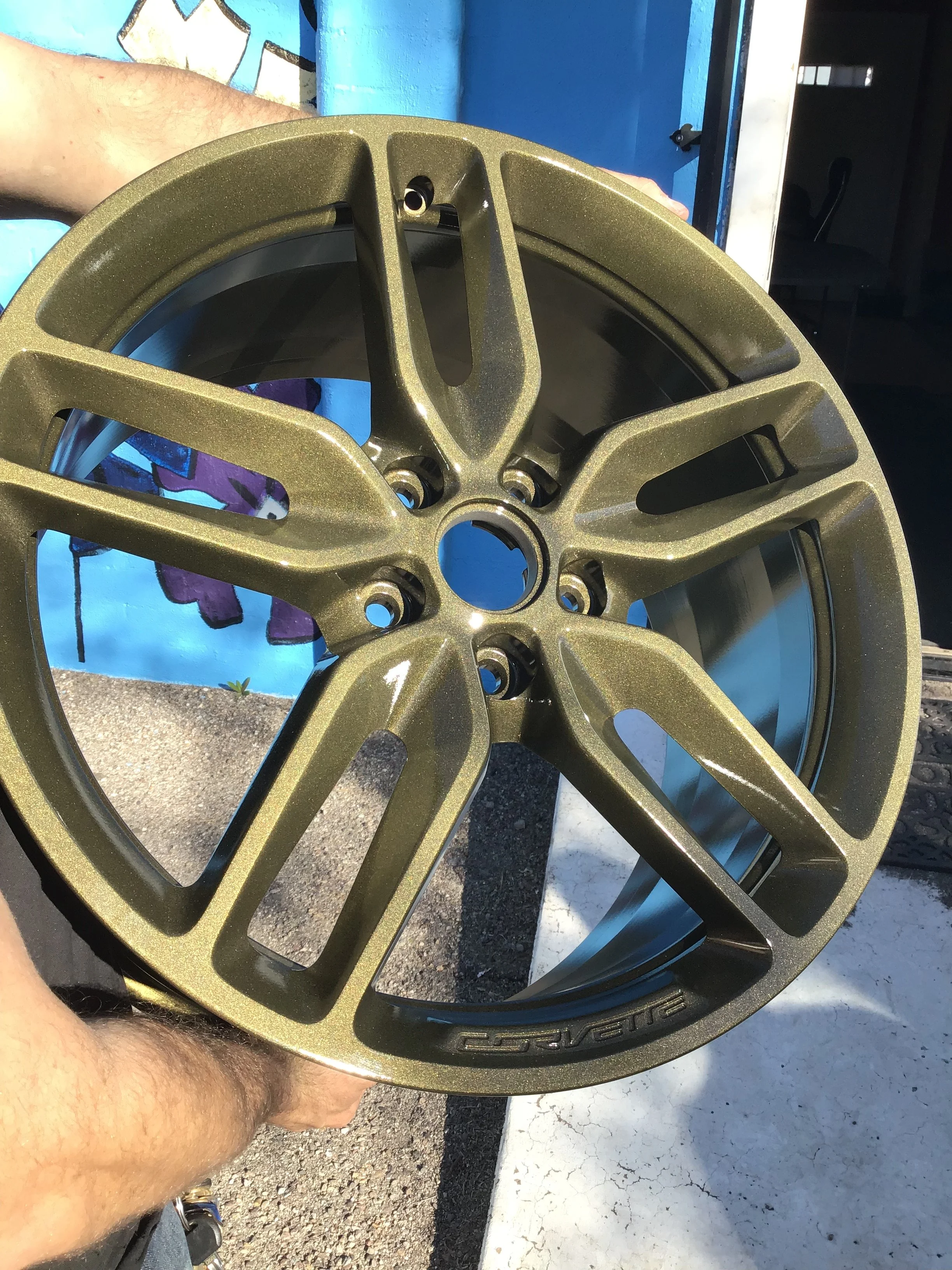 Close-up of a bronze-colored alloy wheel rim being held outdoors, with a blue wall and graffiti in the background.
