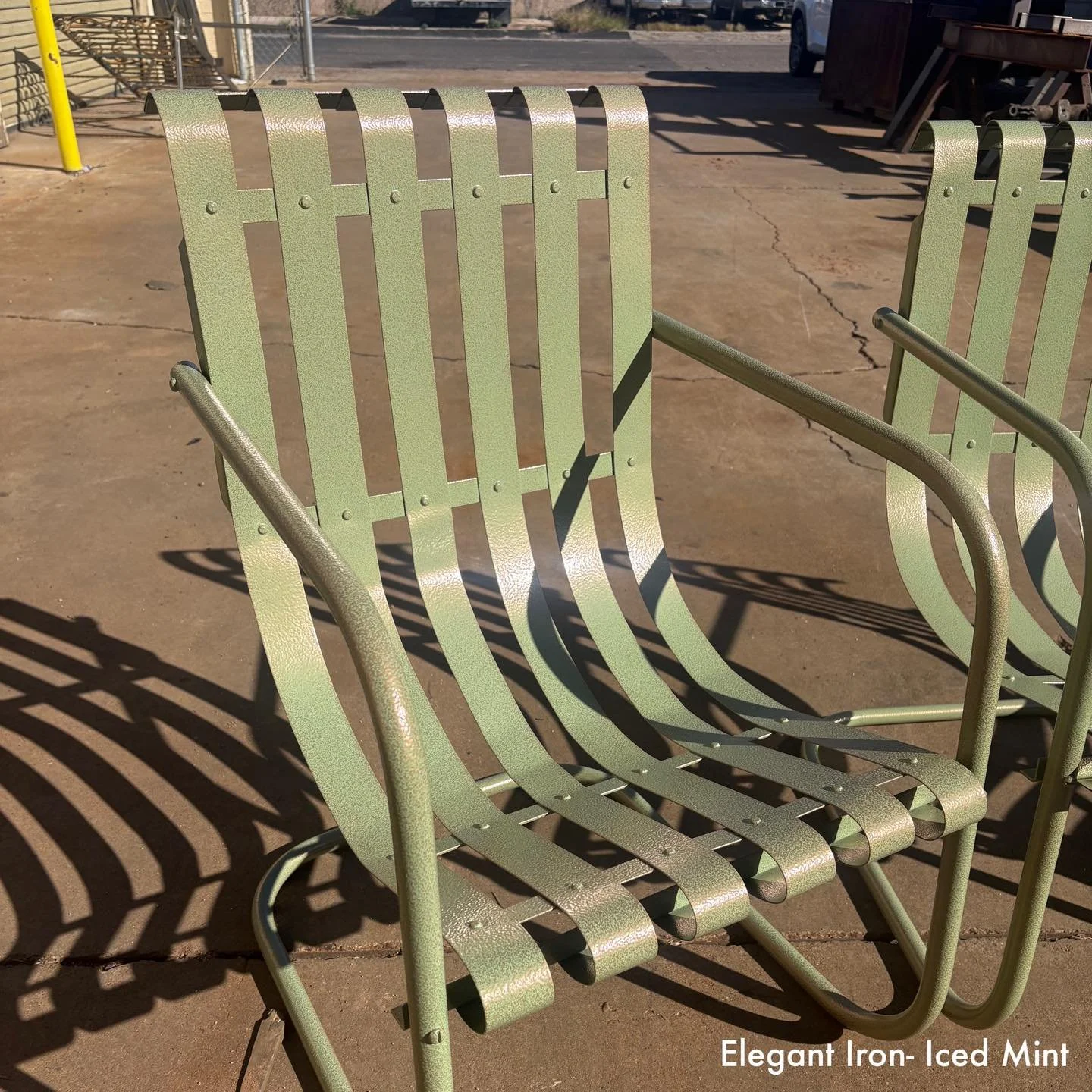 A green metal outdoor chair with a slatted back and seat, casting shadows on a concrete ground. The background features some metal shelves and machinery. The caption reads 'Elegant Iron - Iced Mint'.
