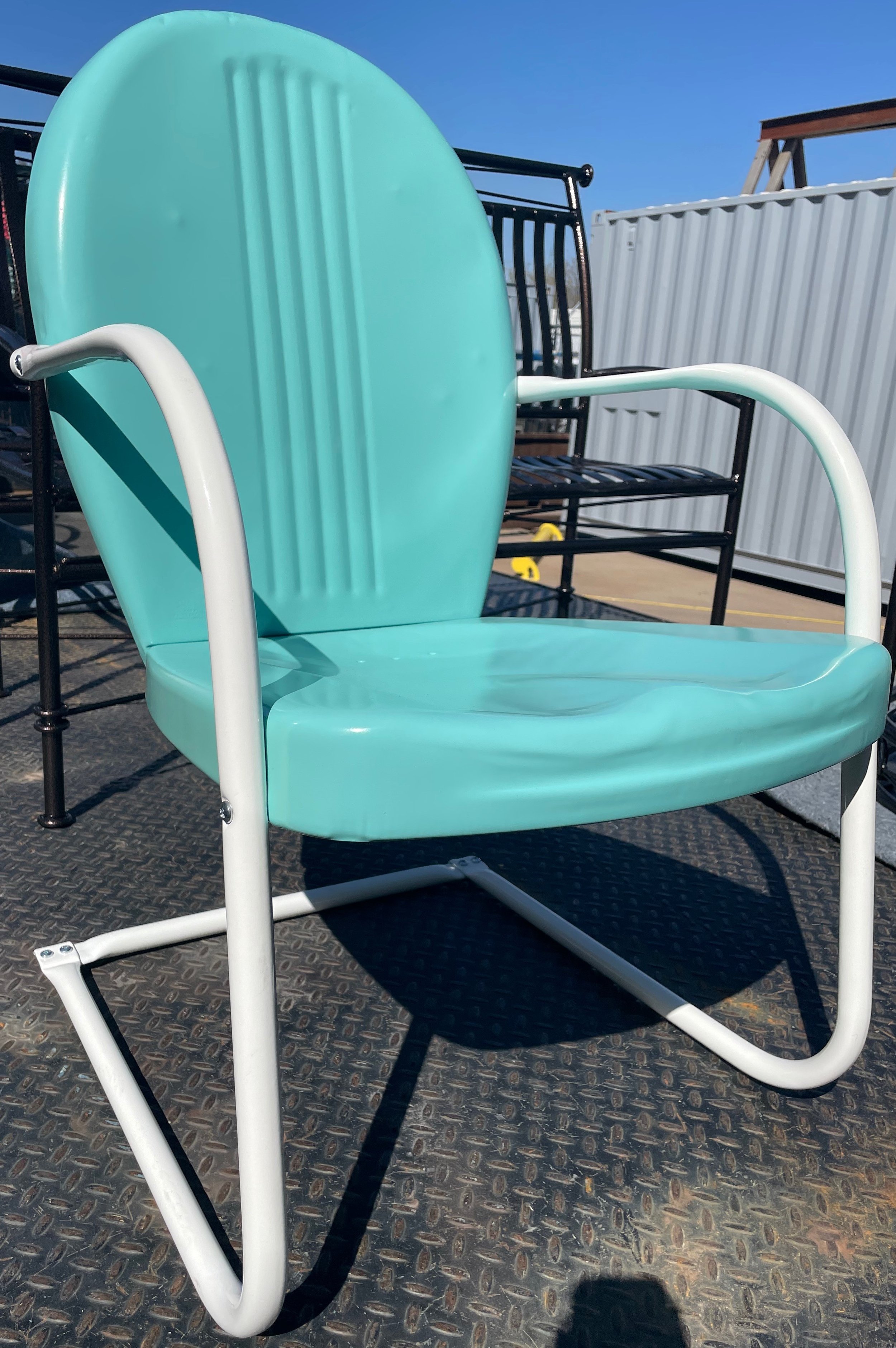 A vintage turquoise metal beach chair with white arms and frame, placed on a textured black surface outdoors, with other black metal chairs and a white fence visible in the background.