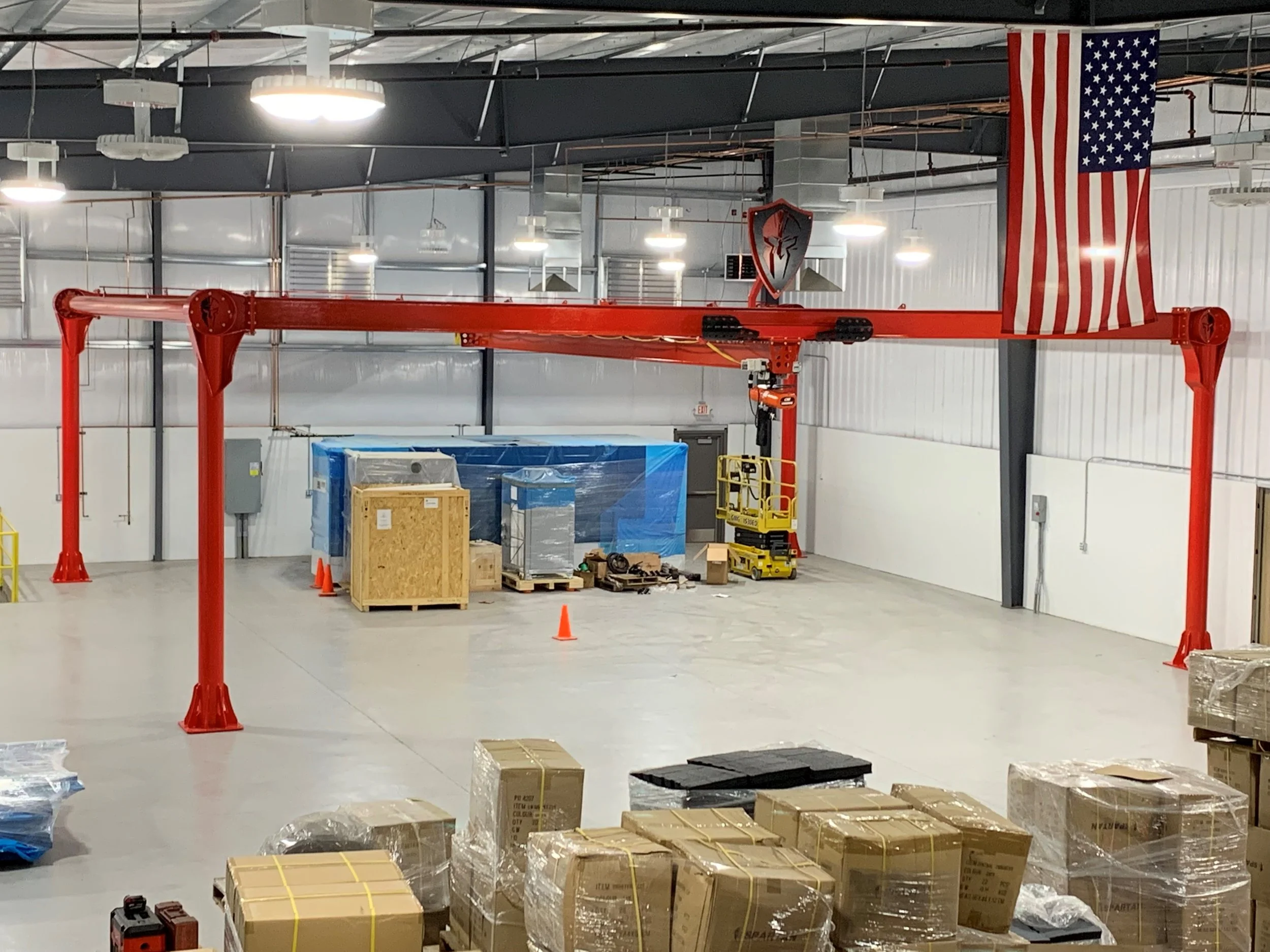 Empty warehouse with packing supplies, pallet stacks, and a large red overhead crane. American flag hangs beside a shield emblem on the wall.