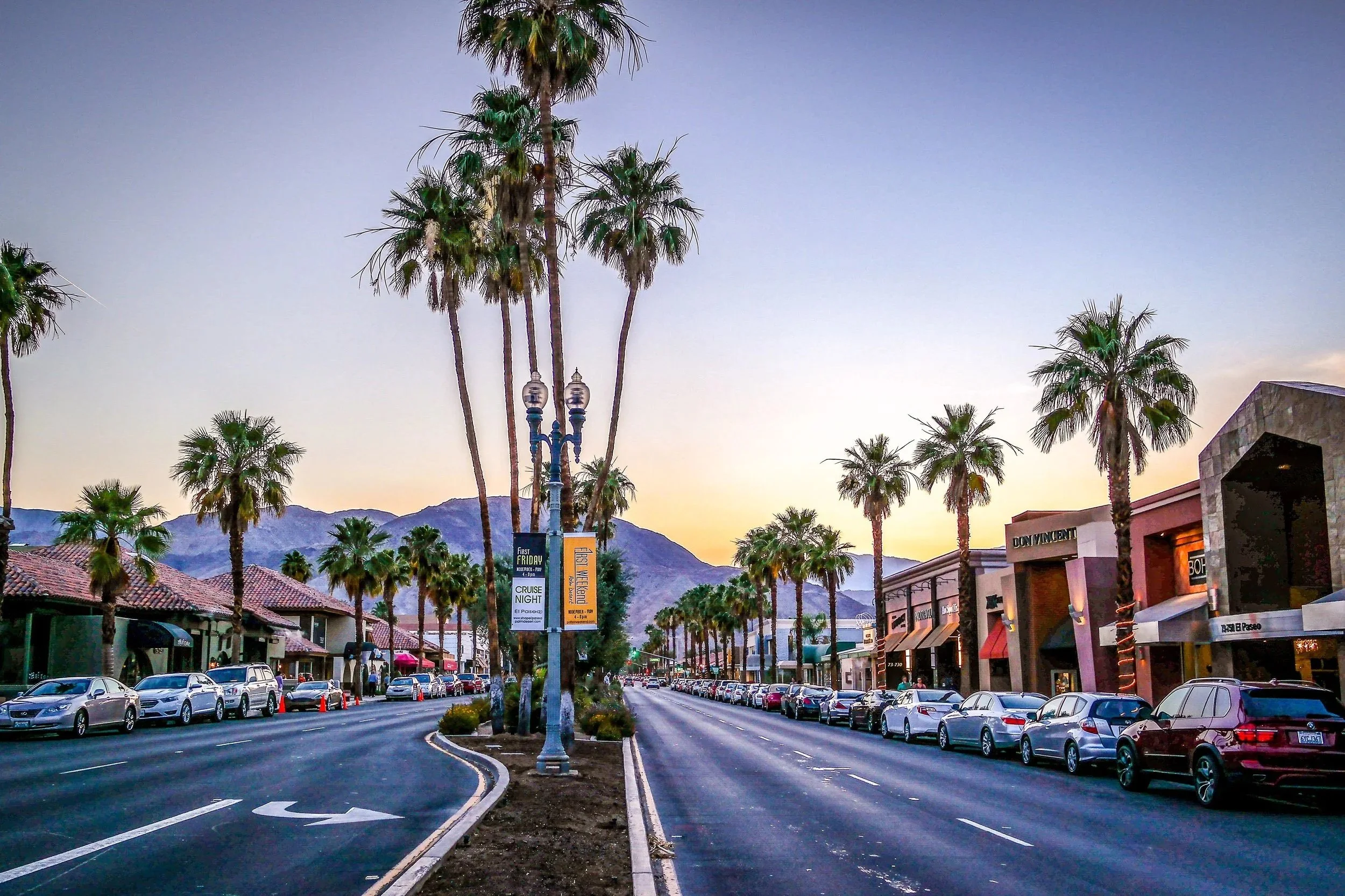 Street with palm trees lining both sides, a row of cars parked along the curb, and buildings with shops and signs. Mountains are visible in the background during sunset.