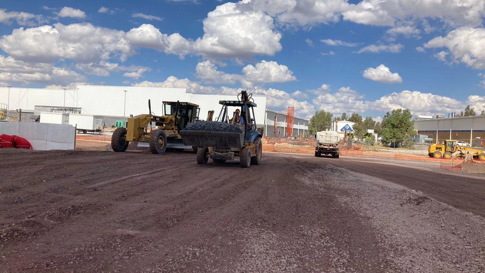 Construction site with bulldozer, dump truck, and other heavy machinery on a dirt lot, with a partly cloudy sky overhead.