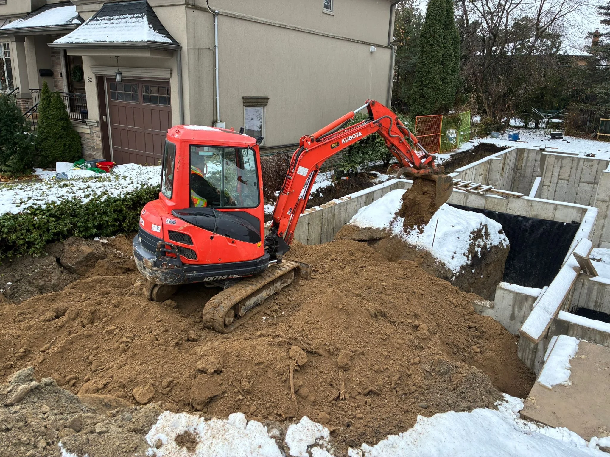 A red Kubota mini excavator digging a large hole in a residential backyard on a snowy day.