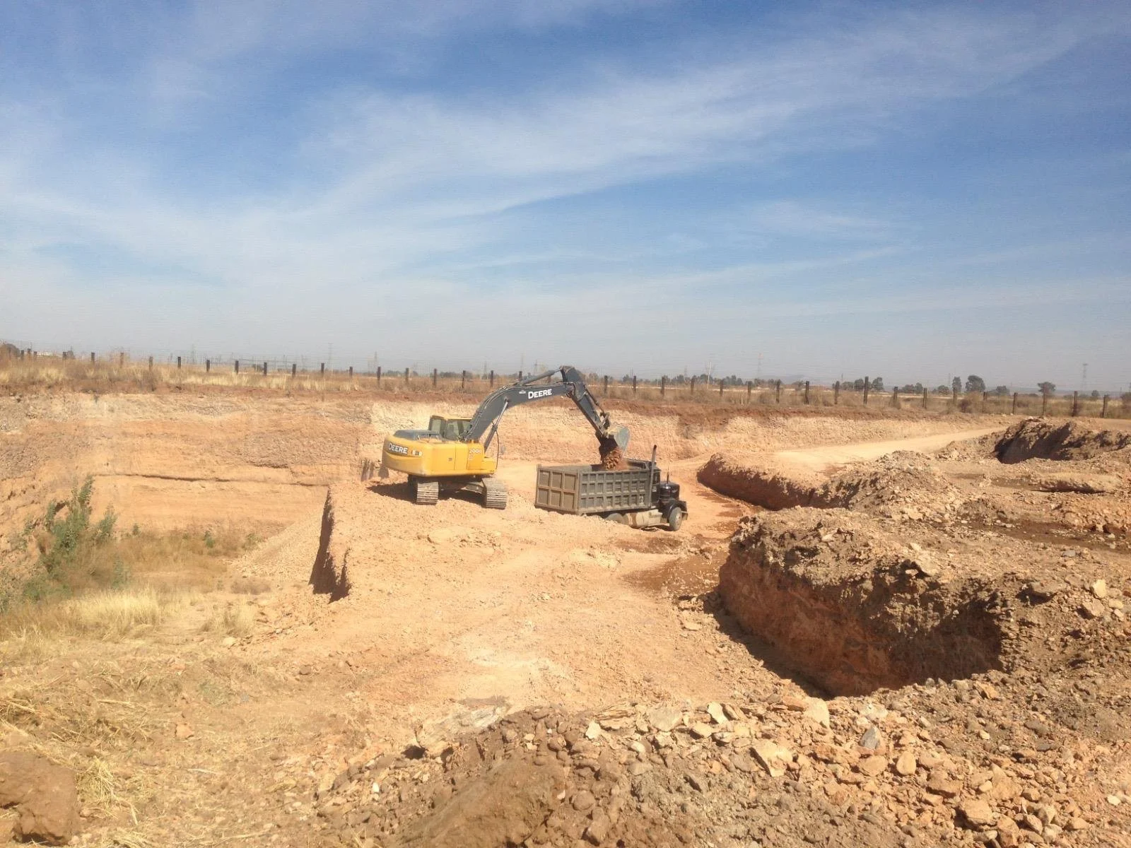 Excavator loading dirt into a dump truck at a construction site in a rural area under a blue sky.