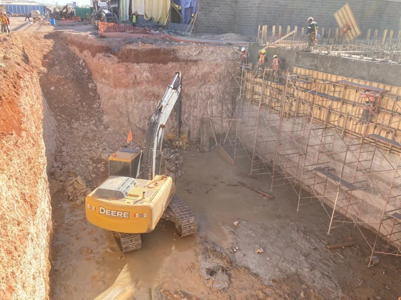 Construction site with workers on scaffolding and a large yellow excavator digging into the ground.