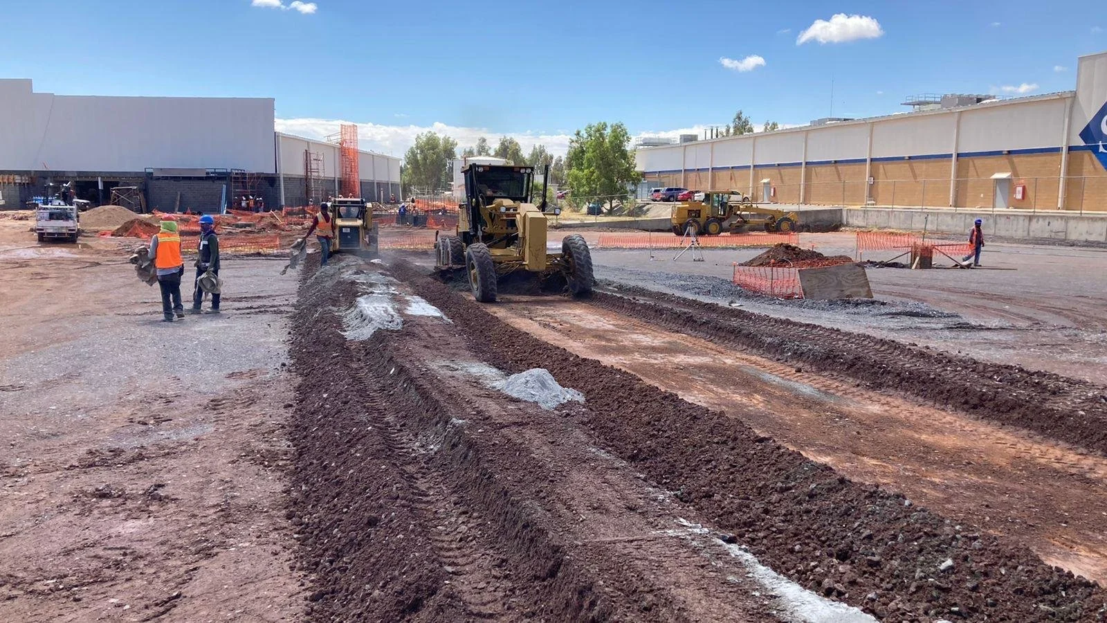 Construction workers leveling and grading soil in a large construction site with heavy machinery, orange safety barriers, and partially built structures in the background.