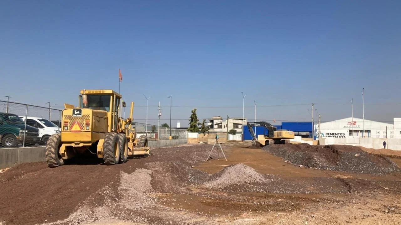 Construction site with yellow bulldozer and excavator, piles of dirt and gravel, cars parked along the side, and commercial buildings in the background under a clear blue sky.