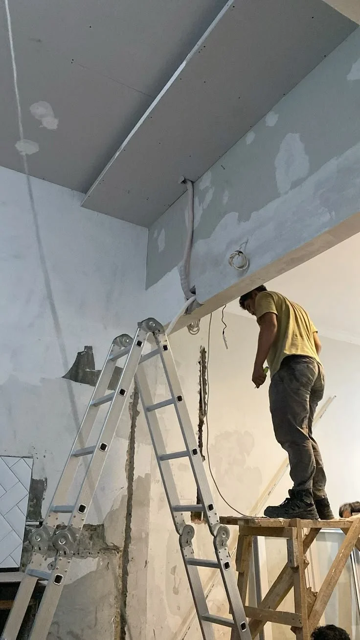 Construction worker standing on a scaffold, working on a ceiling with drywall, surrounded by construction materials and tools.