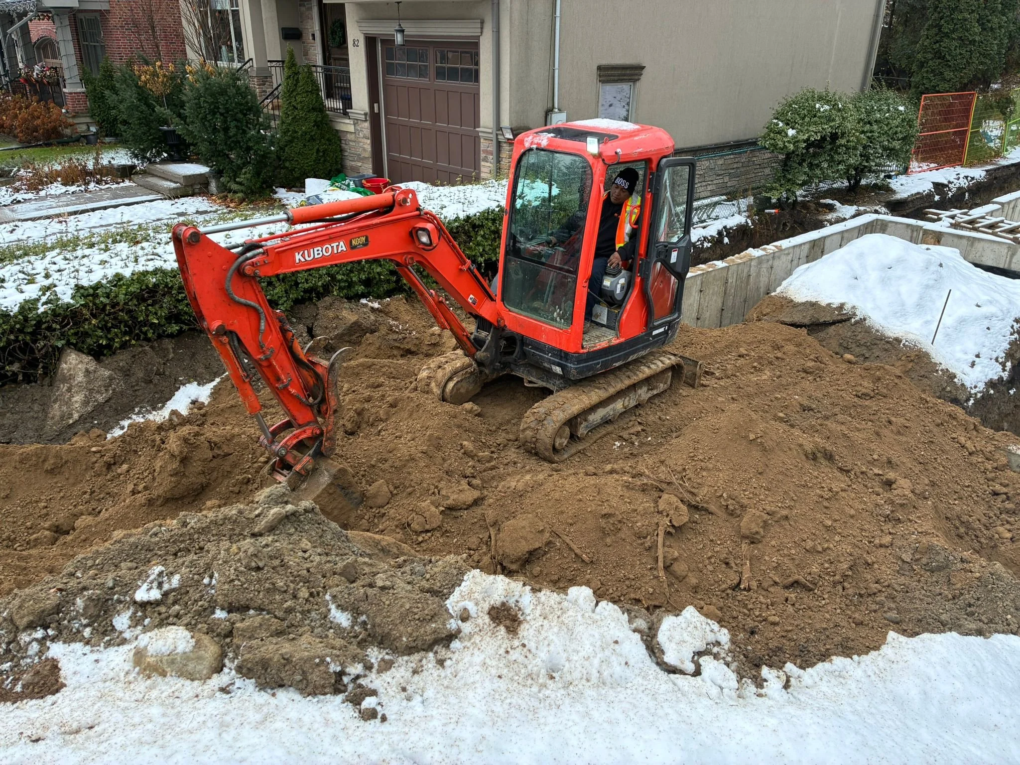 A person operating a small red excavator digging earth on a residential street with snow on the ground.