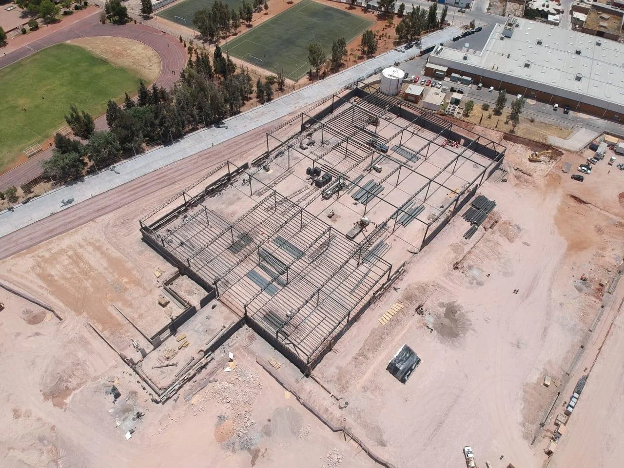 Aerial view of a construction site with steel framework for a building, surrounded by dirt and construction materials, with a nearby school sports field and parking lot.