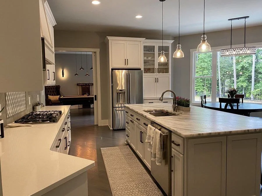 Modern kitchen with white cabinets, stainless steel refrigerator, marble countertops, and pendant lighting, overlooking a dining area with large windows and a view of trees.
